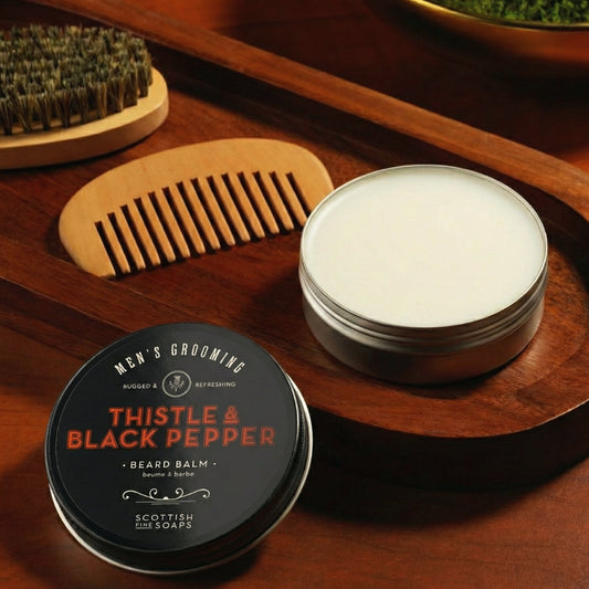 Open Thistle & Black Pepper Beard Balm tub beside a wooden comb and beard brush