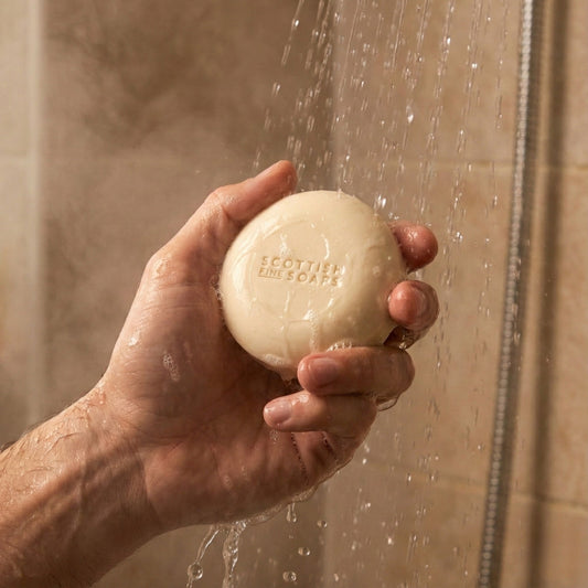 Hand holding a round Thistle & Black Pepper Shampoo Bar in a shower setting