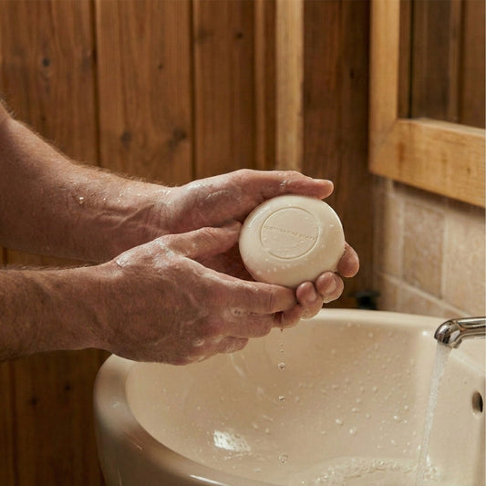 Hands holding a round Thistle & Black Pepper Face & Beard Soap bar over a basin