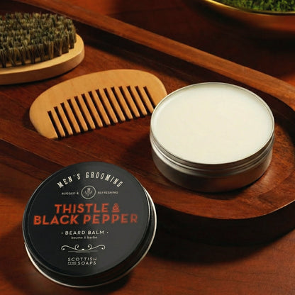 Open Thistle & Black Pepper Beard Balm tub beside a wooden comb and beard brush