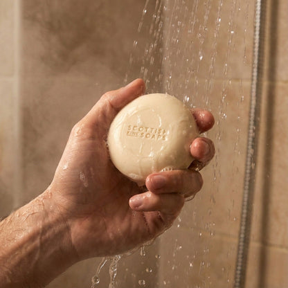 Hand holding a round Thistle & Black Pepper Shampoo Bar in a shower setting