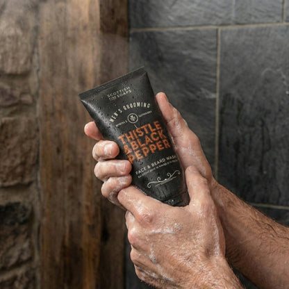 Hand holding Thistle & Black Pepper Face & Beard Wash in a shower setting