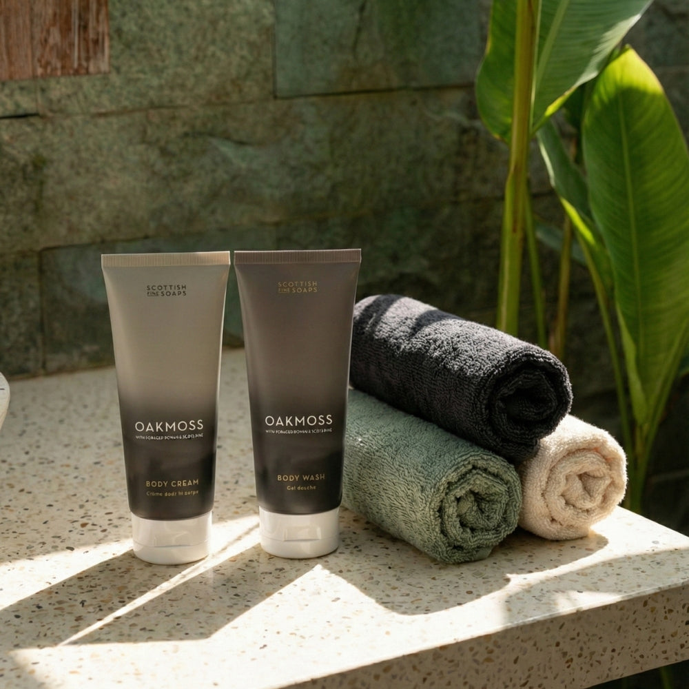 Oakmoss Body Wash and Body Cream tubes beside rolled towels on a bathroom ledge