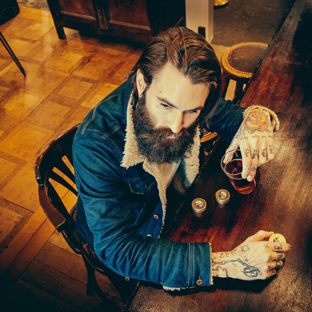 Man with tattoos sitting at a wooden table with a drink, wearing a blue jacket.