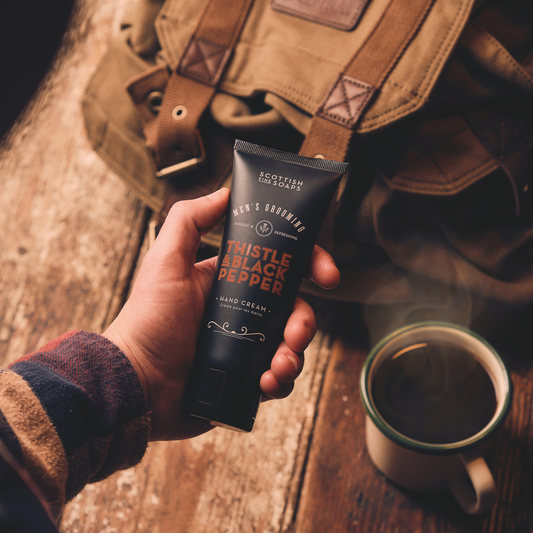 Hand holding Thistle & Black Pepper Hand Cream beside a leather bag and mug